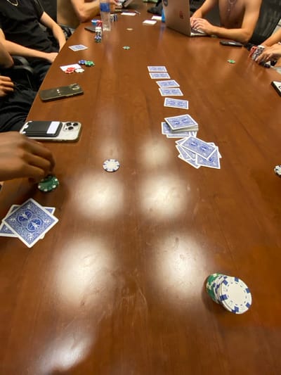 The image depicts an indoor scene located in the City of Albany, United States. A long, dark brown, polished wooden table, displaying multiple reflections from overhead light sources, dominates the frame. At least four individuals are partially visible seated around the table. From the foreground-left, a dark-skinned hand is positioned over a green poker chip, adjacent to three blue-backed playing cards stacked. Two smartphones are on the table in this area: one dark-colored and one silver with a black magnetic wallet attachment. Further along the left, an arm clad in a dark short-sleeved garment is visible. In the mid-background, a shirtless individual wearing a silver neck chain is seated, operating an open silver laptop. To the right, a human hand is present near a stack of red, blue, and green poker chips.
The table is extensively populated with objects. Multiple blue-backed playing cards are arranged in various formations: a linear sequence of single cards, several small stacks, and larger piles. Poker chips are distributed across the table, including individual green, white, and blue chips, as well as a vertical stack of white and blue chips with green underlayers at the bottom-right, and another mixed stack of red, blue, and green chips further up the right side. A clear plastic beverage bottle is situated near the upper-left edge of the table. A dark-colored smartphone is also visible near the individual using the laptop.