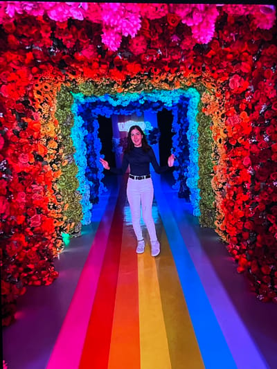 A person is lying down on a rainbow-colored floor in a flower-lined tunnel. The person is wearing a dark long-sleeved shirt, white pants, and white sneakers. They are looking at the camera and smiling. The tunnel walls are covered in a dense arrangement of artificial flowers in various colors, creating a vibrant, multi-hued pathway. The flowers transition through the colors of the rainbow, starting with reds and oranges on the sides, moving to yellows, greens, blues, and purples towards the center. The rainbow floor complements the floral display. The scene appears to be indoors, likely an art installation or a themed attraction. The lighting is bright and colorful, contributing to a cheerful and whimsical atmosphere. There are no clear indications of the time of day or weather. The image quality is slightly blurred, and there is some visible digital noise. No visible text is clearly discernible in the image.