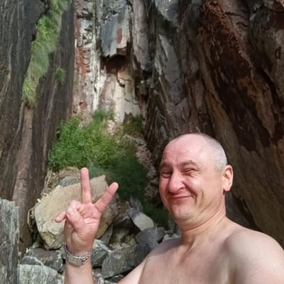 A middle-aged man with a shaved head and a slight smile winks at the camera, holding up two fingers in a peace sign with his left hand. He is shirtless, wearing a silver watch on his left wrist. The man is positioned in the foreground of a rugged, natural environment, with large rock formations and sparse green vegetation behind him. The rocky cliffs suggest a gorge or canyon. The image was taken outdoors during daylight hours, indicated by the natural light illuminating the scene. The location is specified as Ribadedeva, Spain, suggesting a potentially scenic or recreational area within the municipality.