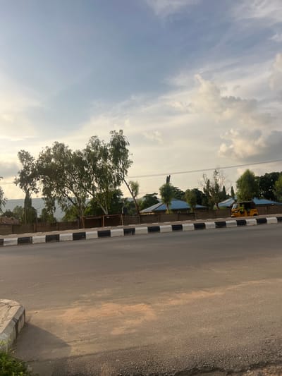 The image depicts a street scene in Kporva, Nigeria. The orientation of the photo is vertical, with the road occupying the right side of the frame and the sky on the left. The road surface appears to be asphalt, with a rougher, gravelly shoulder on the far right. A black and white striped curb runs along the edge of the road, separating it from the sidewalk and buildings.

On the left side of the road, there are several structures, likely houses or shops, with blue roofs. Vegetation, including trees and bushes, is visible behind these buildings. In the distance, a hilly landscape can be seen. A yellow vehicle, possibly a bus or a van, is parked near one of the buildings, along with what looks like a motorcycle. Overhead, power lines stretch across the frame.

The sky is filled with clouds, some appearing white and fluffy, while others are darker and more diffused, suggesting a partly cloudy or overcast day. The lighting indicates it is either early morning or late afternoon, as the sun is casting a soft glow. There is no visible text in the image. The scene appears to be a typical street in a residential or commercial area, with no specific activity or event taking place, other than the general presence of parked vehicles and buildings.