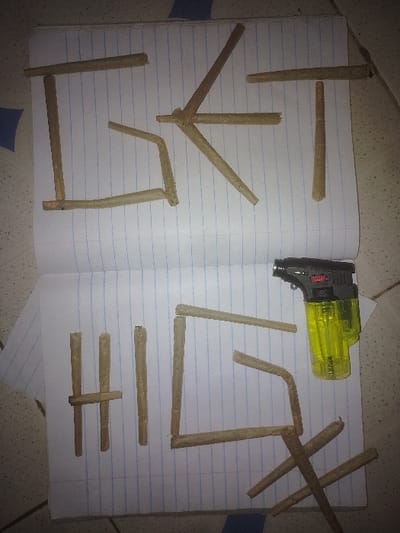 The scene depicts an open, spiral-bound notebook with lined paper placed on a light-colored surface, likely a tiled floor or countertop, in Bamburi ward, Kenya. No individuals are present.
**Subjects/Objects:**
1. **Hand-rolled items:** Numerous cylindrical, hand-rolled items, consistent in appearance with cigarettes or cannabis joints, are arranged on the notebook pages. These items are light brown in color.
2. **Notebook:** An open, standard spiral-bound notebook with blue horizontal lines and a red vertical margin line. The left page is the top page, and the right page is the bottom page of the open spread.
3. **Butane Torch Lighter:** A black and neon-yellow butane torch lighter is positioned on the bottom right page of the notebook. It features a transparent yellow fuel tank and a black top housing the ignition mechanism and flame nozzle.
**Actions/Interactions:**
The hand-rolled items are meticulously arranged to form letters on both pages of the notebook.
1. **Top page:** The items spell the word "ISKT".
2. **Bottom page:** The items spell the letters "H", "I", "L", followed by a complex arrangement of five items forming a stylized character or symbol, which includes a prominent 'X' shape. The butane torch lighter is placed adjacent to this arrangement.
**Environment:**
The background surface is light-colored, possibly off-white or light beige, with subtle darker specks or patterns suggesting a tiled or laminate surface. A small dark blue square is visible on the surface above the notebook.
