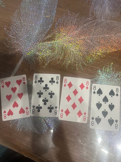 Four playing cards, each displaying the number '8' and a different suit (hearts, clubs, diamonds, and spades), are laid out on a wooden table. The table is adorned with a decorative, glittery design resembling feathers, which reflects light in a prismatic manner. The cards are arranged in a horizontal line from left to right. No individuals are present in the image. The scene appears to be indoors, possibly in a home or gaming area. The provided context of Rosenovo, Bulgaria, does not appear to be directly observable from the image itself, as there are no identifiable landmarks or unique environmental features that would indicate this specific location.