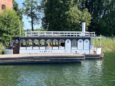 The image shows a white boat named "AKMENA" docked at a wooden pier on a body of water, likely a lake in Trakai, Lithuania. The boat has a dark-colored upper deck with arched windows and a white lower hull. Several passengers are visible inside the boat, seated and looking out. The background features lush green trees, a tall brick tower to the left, and a blue sky. The water is green and slightly rippled. The pier is made of dark wooden planks.