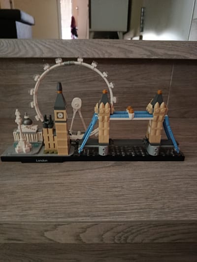 The media file displays a Lego Architecture skyline model depicting several iconic London landmarks. The model is presented on a light brown wooden surface with a distinct wood grain pattern.
The Lego model, arranged on a black base plate, features from left to right: a white monument resembling Nelson's Column and part of Trafalgar Square, the tan-colored Elizabeth Tower (Big Ben) with a clock face, a large white Ferris wheel representing the London Eye, and finally, the Tower Bridge with its tan towers, blue suspension cables, and bridge deck. A small white vehicle is visible near the base of the London Eye structure.
In the blurred background, an indoor setting is visible, including what appears to be the arm of a dark sofa or chair on the left, and a doorway with a light-colored interior beyond it on the right.
There is no activity taking place as it is a static display of an inanimate object. No people are visible. The time of day is not discernible, but the lighting suggests an indoor setting, possibly with natural light from the doorway or artificial lighting.
The visible text "London" is clearly printed in white on the black base of the Lego model. The photograph was taken in Águilas, Spain.