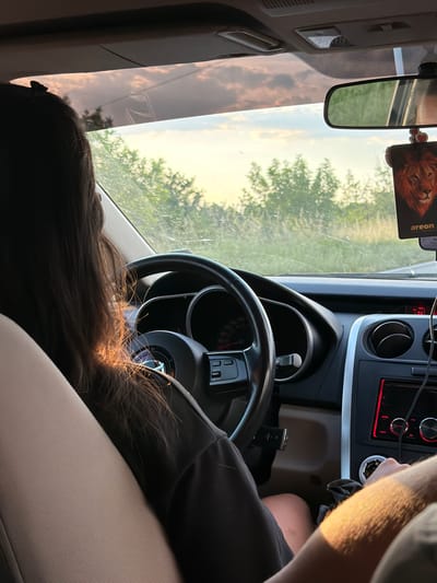 The interior of a car is depicted. A person with long, dark hair is sitting in the passenger seat. The driver's hands are visible, and they appear to be interacting with the car's dashboard. A rear-view mirror hangs above. The car is moving along a road that passes Byal Izvor, Bulgaria, as can be seen through the windshield. The scene is bathed in natural light.