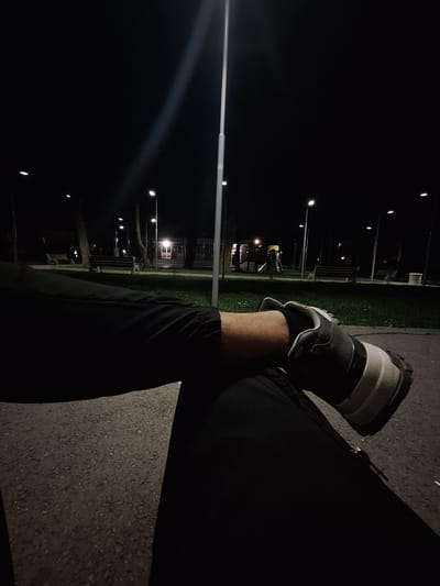 The image is taken at night in an outdoor setting, likely a park or recreation area, indicated by the presence of street lights, benches, and what appears to be playground equipment in the background. The location appears to be in Dolni Chiflik, Bulgaria.
The primary subject is a person wearing dark-colored sweatpants and running shoes. One leg is visible in the foreground, with the running shoe prominently displayed. The person seems to be sitting or resting with their legs crossed.
The environment is dark, with only artificial light illuminating the scene. The ground appears to be paved, possibly asphalt or concrete. There are trees and grass visible in the background, suggesting a green space adjacent to the paved area. The overall impression is one of a quiet or deserted park at night.