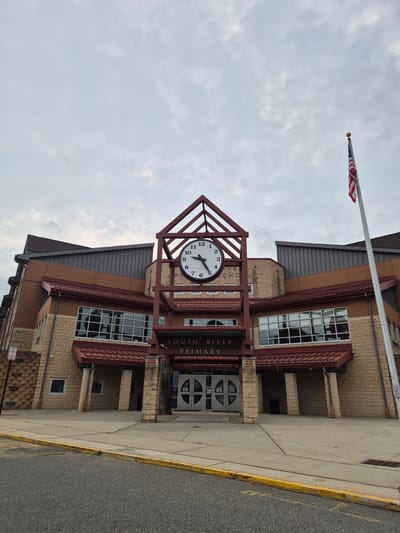 The image shows the front entrance of South River Primary School, located in South River, New Jersey, United States of America. The building features a brick facade with stone accents and a prominent clock above the main entrance. An American flag flies from a flagpole to the right of the building. The sky is overcast with scattered clouds. A sidewalk and street are visible in the foreground. There are no people in the image.