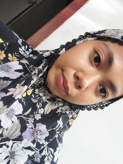 The media file is a close-up, self-taken photograph of a young woman wearing a floral hijab. The woman's face is the primary subject, with her eyes looking directly at the camera. Her expression appears neutral. She is wearing a dark hijab with a black lace trim and a floral pattern featuring various flowers in shades of white, purple, and yellow. The background is a plain, textured white wall, with a hint of a reddish-brown wooden frame on the left side of the image. The lighting suggests an indoor setting, possibly during the daytime. There is no visible text in the image. The overall impression is a personal portrait, likely taken in a domestic setting in Watampone, Indonesia, given the location context.