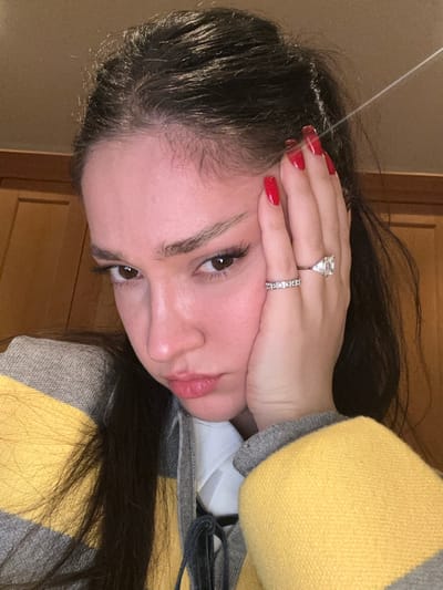 A woman with long dark hair and light skin poses close to the camera. She wears a yellow and gray sweater. She rests her head on her hand, showing off red nail polish and two rings on her finger. She has brown eyes and full lips. The background is a plain, neutral color.