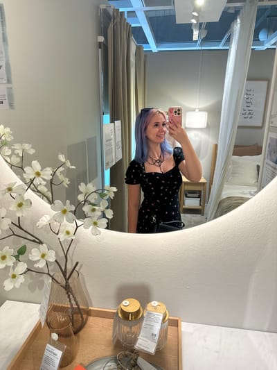 A young woman with dyed blue and pink hair takes a selfie in a large, oval mirror. She is wearing a black dress with a floral pattern, a silver necklace, and sunglasses pushed up on her head. Her phone is pink with a patterned case.

The mirror reflects a room that appears to be a showroom. To the right of the woman, a bed with white and beige linens is visible, covered by a sheer canopy. A small wooden nightstand with books sits beside the bed. In the foreground of the mirror's reflection, there are decorative items on a light-colored surface.

In the lower part of the original image, in front of the mirror, a wooden tray holds two amber-colored glass vases, one larger than the other, with artificial white flowers in the larger vase. There are also two glass jars with gold lids and price tags. The floor appears to be a white marble pattern. The background shows light-colored walls and a ceiling with track lighting.

This scene is located at an IKEA store in the city of Siero, Spain.