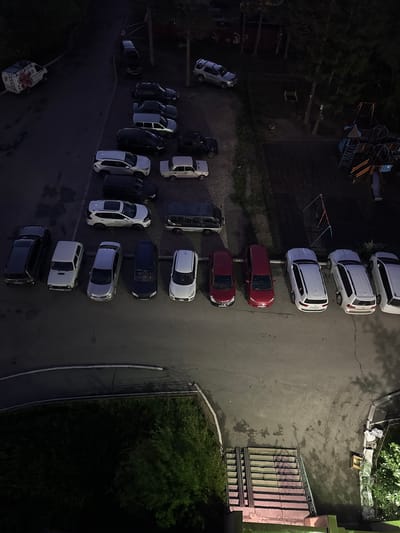 This overhead, nighttime drone shot captures a dimly lit outdoor scene in what appears to be a residential area in Neryungri, Russia. The primary subjects are numerous parked cars, mostly SUVs and sedans, occupying a paved lot that transitions to grass and trees on the left. The cars are arranged in rows and haphazardly, suggesting an informal parking situation.
Several structures are visible, including what looks like a playground with blue metal framing and slides in the upper left quadrant. Further down, there's a gated entrance with a staircase leading up to a building with green trim. The ground is a mix of asphalt and concrete, with some wet patches reflecting the ambient light. Shadows are cast by the cars and trees, indicating artificial lighting or residual daylight.
The time of day is clearly night, given the dark sky and the artificial illumination. The weather appears calm, with no indication of rain or strong winds. The scene evokes a sense of quiet suburban or apartment complex life at night. There are no visible people or animals. No text is discernible in the image.