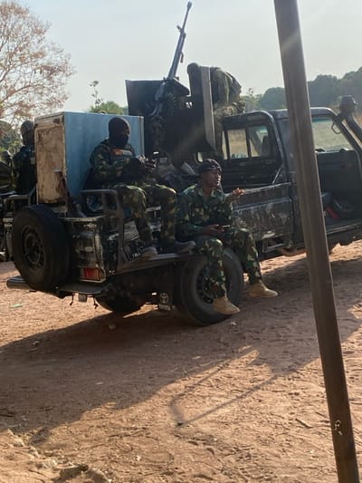 The image shows a military vehicle, a Toyota pickup truck, with soldiers in camouflage uniforms riding in the back. Some soldiers are lying down, while others are seated. A heavy machine gun is mounted on the vehicle. The setting appears to be outdoors, possibly in a rural or desert-like area, with dusty ground.