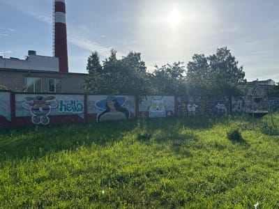 The image depicts an outdoor scene with a graffiti-covered concrete wall in the foreground, set against a backdrop of trees, a building, and a smokestack. The wall features various cartoonish murals, including a character with large ears and rainbow stripes, the word "hello," a woman with a wide-brimmed hat, and a stylized cat. Beyond the wall, lush green grass covers the ground. In the background, a tall, red and white striped smokestack rises into a bright, sunny sky with scattered clouds. A low-rise brick building is visible to the left of the smokestack. Trees with full green foliage form a dense line behind the wall. To the right, a portion of a metal play structure or garden feature is visible. The overall setting appears to be an urban or industrial area with a touch of greenery.