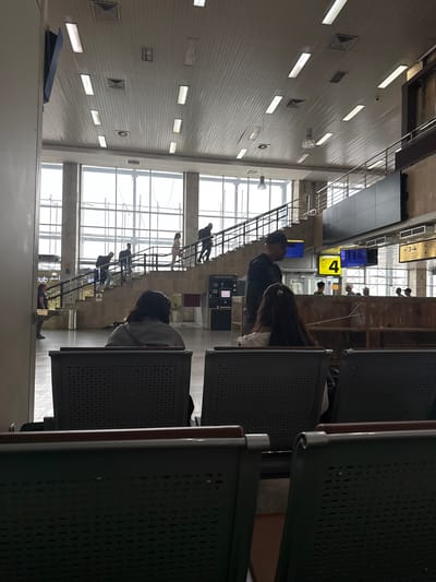 The image depicts the interior of an airport terminal, possibly at Manas International Airport in Bishkek, Kyrgyzstan, judging by the available visual cues. The scene is characterized by a large, open space with a high, paneled ceiling featuring numerous fluorescent light fixtures and air vents. A prominent staircase with metal railings ascends to the right, with several individuals walking up it. In the foreground, rows of dark, perforated waiting chairs are visible, with two individuals seated facing away from the camera in the lower half of the frame. Further back, a yellow digital sign displaying the number "4" is mounted on a wall, suggesting a gate or zone number. Other digital displays and signs, some with Cyrillic text, are also present. People are scattered throughout the terminal, some walking, others possibly waiting. The overall atmosphere suggests a functional, public transit hub.