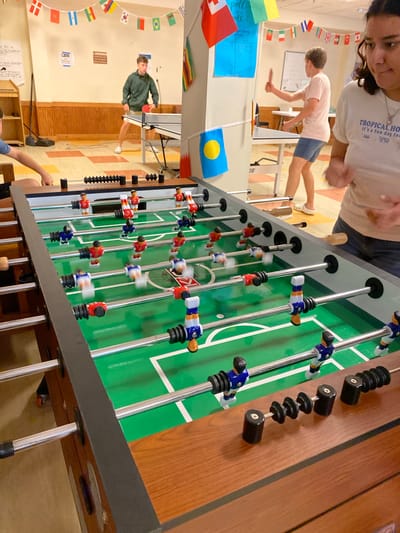 In Washington Township, USA, a foosball table dominates the foreground of an indoor recreation area. The table features a green playing surface with white markings and rows of miniature red, blue, and white player figures on metal rods. In the background, two young people are engaged in a table tennis match. One, wearing a green hooded jacket and dark shorts, stands to the left of a table tennis table, holding a red paddle. The other, in a pink t-shirt and blue shorts, is on the right, also with a paddle. The room is decorated with a string of international flags and various posters on the walls. The floor has a checkerboard pattern of orange and yellow tiles.