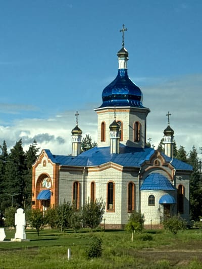 The image displays an Orthodox church located in Kapitanivka, Ukraine. The church building features light-toned brick walls with accents of reddish-brown brick outlining arched windows and decorative elements. It has a central, prominent dome and four smaller domes, all covered in a reflective metallic blue material. Each dome is topped with a golden, onion-shaped element surmounted by an Orthodox cross; the central cross exhibits greater ornamentation. The main roof sections are also bright metallic blue. An arched entrance on the left facade includes an overhead fresco and a small blue canopy. The structure stands on a manicured green lawn containing small shrubs and deciduous trees. In the left foreground, a white, monolithic cross-shaped monument is visible. A dense line of dark green coniferous trees forms the background behind the church. The sky is partially cloudy blue. No individuals are present in the scene.