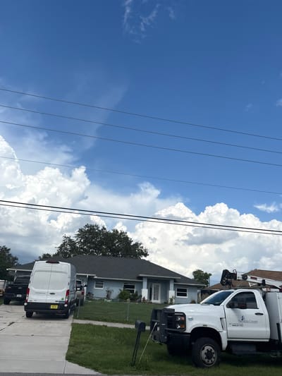 The image depicts a residential street scene in North Port, Florida, USA. In the foreground, a white utility truck with a crane attached is parked on the grass. The truck has "Sarasota County" and "Silverado 6600 HD" written on its side. To the left of the truck, a white cargo van is parked on a concrete driveway. Behind the van, a black pickup truck is partially visible. The background features a light blue single-story house with a dark roof, surrounded by a green lawn and trees. Power lines are strung across the sky, which is predominantly blue with large, fluffy white clouds. The overall impression is a suburban setting on a bright day, possibly with ongoing work or services taking place due to the presence of the utility vehicles.