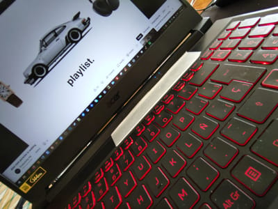 The image shows a close-up of an open Acer laptop. The screen displays a YouTube playlist with a white Porsche and the word "playlist." Red backlighting illuminates the black keys of the keyboard, which are in a Spanish keyboard layout. A 144Hz Full HD sticker is visible on the laptop's lower left. There are no people visible, and the location is not specified. The environment is unclear, although there is a wooden surface visible near the laptop. The essential objects are a laptop, keyboard, and the YouTube interface.