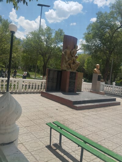 The image captures a monument in Naryn City, Kyrgyzstan, dedicated to fallen soldiers, likely from World War II. The monument features a central bronze sculpture of several figures, set against a dark, curved plaque inscribed with names. To the right of this, a bust of a man in uniform stands on a pedestal. The monument is situated in a paved plaza with a white, ornate railing in the background. Several trees with lush green foliage are visible, suggesting a park or public garden setting. The sky is blue with scattered white clouds. In the foreground, a green park bench sits on the patterned paving stones. A lamppost with a spherical light fixture is visible to the left of the monument. A few individuals are present in the middle ground, near the railing, suggesting the site is accessible to the public. The overall scene is one of a well-maintained public space honoring historical remembrance.