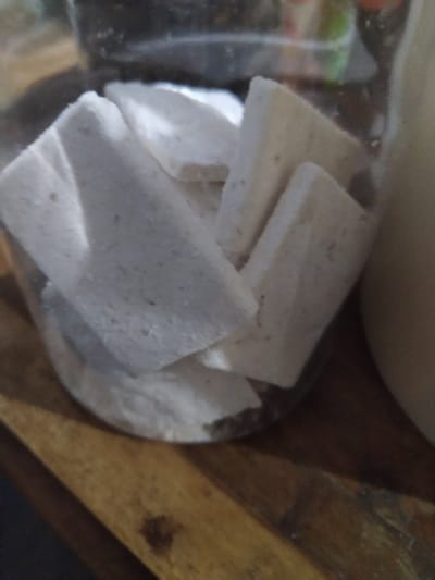 The image presents a close-up view of several rectangular, off-white objects contained within a transparent plastic bag or container. These objects, possibly food items, are consistently block-shaped, though some display slightly irregular or crumbly edges. Their surfaces appear dry and textured, with visible small, dark speckles embedded throughout. A fine, light-colored, powdery residue, similar in color to the objects themselves, is noticeable at the bottom of the container and adhering to the objects, suggesting a starch or flour coating.

In the background, a wooden surface with a visible grain and some darker marks is present to the right and bottom right. A blurry, light-colored, undefined object is visible in the top background. The scene appears to be indoors.

There is no activity taking place; the objects are static, likely being stored or prepared. No visible text is present in the image. Given the visual characteristics and the location context of Gampongbaro, Indonesia, these objects strongly resemble uncooked crackers (kerupuk mentah) or a similar type of dried, starch-based snack or ingredient, typically intended for further preparation like frying. The time of day, weather, or emotions cannot be determined from the image.