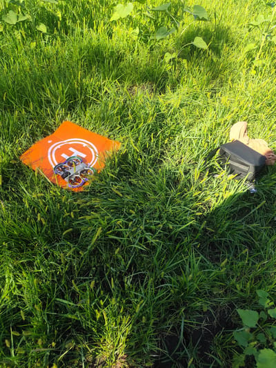 In Bozhurishte, Bulgaria, a quadcopter drone rests on an orange landing pad in a grassy area. Adjacent to the drone, a black carrying case and a tan piece of clothing are visible. Tall plants, possibly sunflowers, and dense green grass populate the background, illuminated by sunlight.