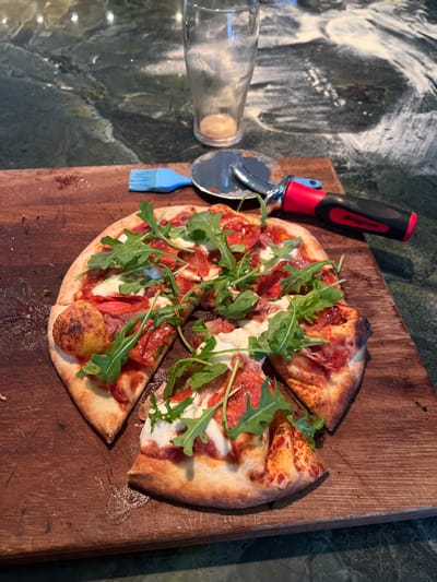 An overhead view captures a freshly sliced pizza on a well-used wooden cutting board, resting on a dark green, veined stone countertop. The pizza features a golden-brown crust and is topped with red tomato sauce, melted white cheese (likely mozzarella or burrata), slices of cured red meat (possibly pepperoni or salami), and fresh green arugula leaves. One large irregular slice has been separated from the main pizza. To the upper right of the pizza, a silver pizza cutter with a red and black "Snap-on" branded handle lies beside a small blue silicone basting brush. In the background, an empty, clear glass tumbler stands on the countertop, which is visibly dusted with white flour, suggesting recent food preparation. No individuals are present. The precise location within Hornsea, United Kingdom, cannot be determined from the visual information provided.