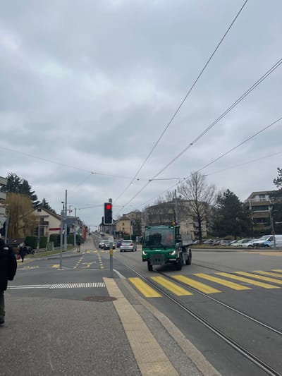 This image captures a street scene in Meyrin, Switzerland, on an overcast day. The street, which features tram tracks and yellow pedestrian crossing stripes, extends from the foreground into the distance towards the right. A prominent green utility vehicle, resembling a small road maintenance or tractor-like vehicle, is visible on the road, positioned near the tram tracks and pedestrian crossing. It appears to be stopped, facing generally towards the right side of the frame.

A red traffic light is clearly visible on the left side of the image, indicating that traffic is halted. Several other cars are visible further down the street, some parked and others seemingly stopped in traffic. Residential or commercial buildings line both sides of the road, with some bare trees suggesting a late autumn or winter season. Overhead electrical wires, likely for trams or trolleybuses, crisscross the grey, cloudy sky. In the bottom right corner, part of a person in dark clothing is visible, possibly the photographer or a passerby. Road markings include white dashed lines and yellow symbols, possibly indicating a bicycle lane. No specific text is clearly legible in the image.