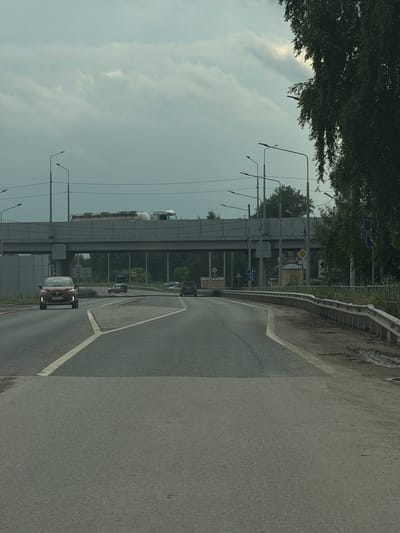 The image captures a view from a moving vehicle on a multi-lane road under an overcast sky. A concrete overpass with a white railing is visible in the mid-ground, with a large white truck carrying a tanker driving across it. Several streetlights are positioned along the road and overpass. On the right side of the road, a metal guardrail runs along a muddy embankment bordered by trees. A red car with its headlights on is approaching in the left lane. Further ahead, several other vehicles are on the road, including a light-colored SUV. The road is marked with white lane lines and a pedestrian crossing area. Signs are visible on the side of the road, including a diamond-shaped yield sign. A small, light-yellow building with a domed roof is situated behind the overpass on the right side. The environment appears to be a suburban or semi-urban area, with vegetation and trees present.