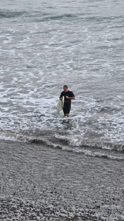 A man in a black wetsuit stands in the ocean in Encinitas, California, holding a white surfboard. The water is choppy and foamy, with waves breaking around his knees. The man is looking down at the surfboard, and appears to be preparing to go surfing. The background is blurred, with the ocean extending to the horizon. The beach in the foreground is covered in pebbles and rocks.