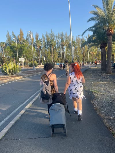 Two individuals dressed in costumes are walking along a street in Campo Internacional, Spain. One person is wearing a leopard-print outfit and the other is wearing a white dress with black spots. The person in leopard print is pulling a gray luggage cart. The scene takes place on a sunny day near palm trees, cactuses, and other tropical vegetation. A street light is visible along the road. Other people are walking in the background.