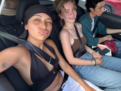 This media file captures a close-up, high-angle view of three people relaxing inside a car, seemingly lying down with their heads oriented towards the bottom of the frame (if viewed upright). The overall atmosphere is casual and relaxed.

In the center, a young woman with reddish-blonde hair is smiling directly at the camera. She wears a brown spaghetti strap top and a silver necklace, with a seatbelt visible across her chest. To her right, a person with a dark skin tone, freckles, and a nose piercing is seen winking and puckering their lips playfully. They wear a black headwrap or durag and a black top or dress with a deep V-neck, adorned with a long gold chain necklace featuring small charms. A lotus flower tattoo is visible on their left arm, and underarm hair is also visible. This individual is also wearing a seatbelt.

To the left of the blonde woman, a third person is partially visible, lying down with their head to the left, wearing a green collared shirt. Further to the top left of the frame (if viewed upright), another person's torso and legs are visible, dressed in light blue jeans and a white t-shirt, wearing bracelets on their wrist. A light scar is present on their forearm.

Various personal items are scattered around: a red fabric item, two smartphones (one black, one silver/grey), and what appears to be a green book or notebook and an orange-covered book, resting between the occupants. The car interior features black seats and seatbelts. Part of a red car and a tire are visible outside the window, suggesting a daytime setting. There is no visible text legible enough to describe.

The scene depicts friends or companions resting during travel in Santa María Colotepec, Mexico.