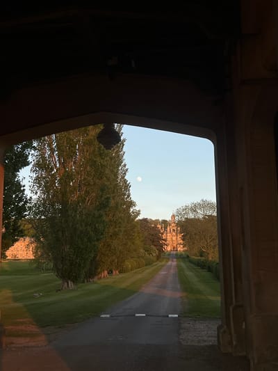 An outdoor scene is captured through the dark, roughly textured archway of a stone or concrete structure, which frames a landscape beyond. The view looks down a long, straight paved driveway or road that recedes into the distance, flanked on both sides by expansive green lawns and numerous mature trees with dense foliage. The road has subtle markings, including a white line on its right edge.
In the mid-distance, a grand, multi-story stone building, likely a historic manor or castle, rises amidst the trees, its façade partially obscured by greenery but illuminated by the soft, warm light of what appears to be dusk or early evening. Its architecture suggests a stately home characteristic of the United Kingdom, specifically fitting the context of South Kesteven. The clear, light blue sky above features a prominent full moon towards the upper left of the framed scene. A dark, round, possibly decorative object or light fixture is visible hanging from the left side of the archway in the foreground.
The lighting creates a serene atmosphere, highlighting the textures of the building and trees. There are no people or animals visible, and no discernible text. The overall scene conveys a sense of tranquility and historical grandeur.