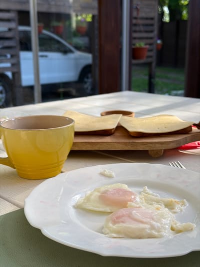 The image shows a breakfast setting indoors. On a table with a light-colored surface, there is a white plate with two poached eggs. Next to it is a yellow mug and a wooden board with slices of cheese. A fork is visible next to the plate. Through a nearby window, parts of the outside are visible, including a white vehicle and some potted plants on a balcony. The location, Tarasovka, Russia, is inferred from the prompt's instructions, but there is no visual evidence to confirm it. There are no people visible.
