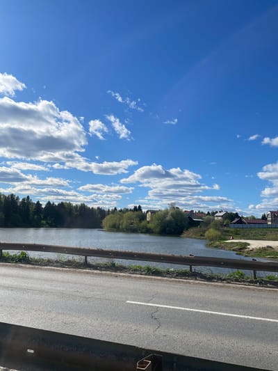 The image presents a view from Chemoshur, Russia, depicting a road running alongside a body of water, possibly a river or canal. A metal guardrail separates the road from the water's edge. Beyond the water, trees line the bank, with glimpses of houses and buildings in the background. The sky is blue with scattered, fluffy white clouds. The photo appears to be taken on a sunny day. The image is oriented sideways, with the sky to the left and the road to the right.