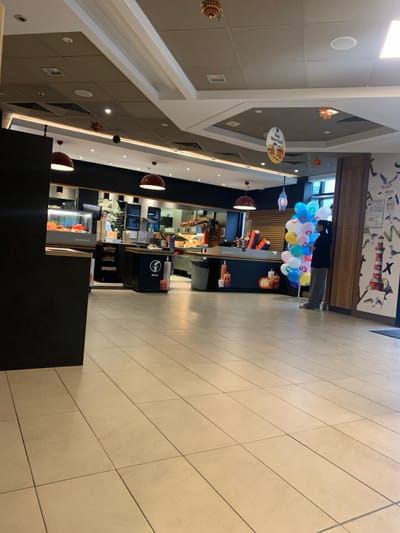 The image displays the interior of a McDonald's restaurant located in Fontenay-sous-Bois, France, viewed from the dining area towards the service counter and kitchen.

The foreground features a clean, light-colored tiled floor. A dark, solid partition wall is present on the far left.

In the midground, the primary service counter stretches across the width of the frame. This counter system is predominantly dark blue and black, with a "SALAD BAR" sign visible on a section to the left. Several dark blue counter units with an "M" logo and orange pumpkin-shaped decorations are positioned along the service line. Behind the counter, a brightly lit kitchen area with stainless steel surfaces and various equipment is visible. One male employee, wearing a blue shirt, is partially visible within the kitchen area.

To the right of the counter, a female individual stands, facing the counter area. She is dressed in a dark jacket and light grey pants. Beside her is a large, vertical cluster of approximately 30-40 helium balloons in various colors, including light blue, yellow, white, and pink.

The right wall features a decorative mural composed of stylized illustrations depicting seagulls, lighthouses, and nautical elements against a light background. A dark entrance mat is visible on the floor near this wall.

The ceiling consists of a multi-layered design with white and grey acoustical tiles, integrating both rectangular and circular light fixtures, along with ventilation grilles. Several red dome-shaped pendant lights are suspended. Decorative elements, including a hanging rectangular sign advertising "Fish New York" and small dark spherical ornaments, are also visible suspended from the ceiling.