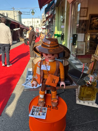 The image captures an outdoor market scene in Saint-Ouen-sur-Seine, France, specifically within the Puces de Saint-Ouen flea market. In the immediate foreground, a large, customized Playmobil-style figure stands centrally on an orange metal barrel marked with "HERMES PARIS" in black text. The figure is dressed in an orange and brown ensemble, featuring a brown wide-brimmed hat inscribed with "HERMES PARIS" and an "H" logo, an orange scarf, brown shoes with "H" buckles, and an orange mini-purse with an "H" logo carried on a gold chain. A small white tag with handwritten text is attached to the figure's neck. To the right of the figure, on a white display stand, two clear glass perfume bottles, one labeled "HERITAGE," are visible.
A vibrant red carpet covers the ground, extending from the foreground into the background, marking a pedestrian pathway. On the left side of the carpet, a man in a light beige jacket and dark pants is walking away from the viewer. Further down the carpet, several other indistinct individuals are walking, including a woman in a dark brown top and pants facing left. The pathway is flanked by commercial establishments with various awnings (red, blue) and display windows, suggesting shops or stalls. The background reveals more market activity and buildings under a clear blue sky. An overhead streetlamp fixture with two lanterns is visible above the street. A shop window to the right, behind the Playmobil figure, displays framed artwork and mirrors, with a dark chair visible inside.