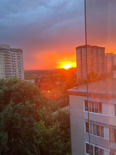 This media file depicts a vivid sunset over an urban landscape in Lytkarino, Russia. The image is oriented vertically, with the horizon running horizontally across the center.
The primary subjects are several apartment buildings and a prominent, bright sunset. In the foreground, on the right side of the image, is a cluster of lush green trees, suggesting a park or residential area with abundant foliage. Behind the trees, a portion of a building with a brick facade and multiple windows is visible. In the mid-ground, there are two large apartment buildings. The one on the left is tall and features a repetitive pattern of balconies and windows, with a facade that transitions from a lighter color at the top to orange tones lower down. The building on the right is partially obscured by the trees and appears to be a lighter color with numerous windows. Beyond these buildings, a sprawling cityscape is visible, stretching towards the horizon.
The activity captured is the natural event of a sunset. The sun is low in the sky, casting a warm, intense orange and yellow glow that dominates the sky and reflects on the surfaces of the buildings and trees. This lighting creates a dramatic and atmospheric scene. The weather appears to be clear, with the sky transitioning from a deep purple-blue on the left to a brilliant orange and yellow towards the sun. The presence of clouds is minimal, allowing the full intensity of the sunset to be visible.
Notable details include the strong lens flare from the sun, visible as a bright, reddish-orange circle in the middle-right of the image. A thin horizontal line, possibly a wire or railing, runs across the middle of the frame, dividing the sky from the cityscape. Tiny specks in the sky above the sun might be birds or insects. The overall mood conveyed is one of warmth, beauty, and the peaceful end of a day. No visible text is present in the image.