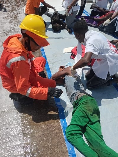 The scene depicts an emergency response situation in Katabu, Nigeria, located at coordinates 10.71 latitude and 7.514 longitude. A man wearing a bright orange jumpsuit and a yellow hard hat is kneeling on the ground. He is looking down and appears to be inspecting something. A second man wearing a white shirt with a red cross on it is kneeling next to a man lying on the ground with a white sheet beneath him. The man in the white shirt is holding a white cloth in his right hand. There is a red first-aid kit nearby. The man on the ground is wearing green pants and a dark blue shirt. Several other people are in the background. The scene is a public area, likely a street or sidewalk, with a white tarp spread out on the ground. The surface of the ground is wet, suggesting there may have been recent rainfall. The scene appears to be a response to a medical emergency.