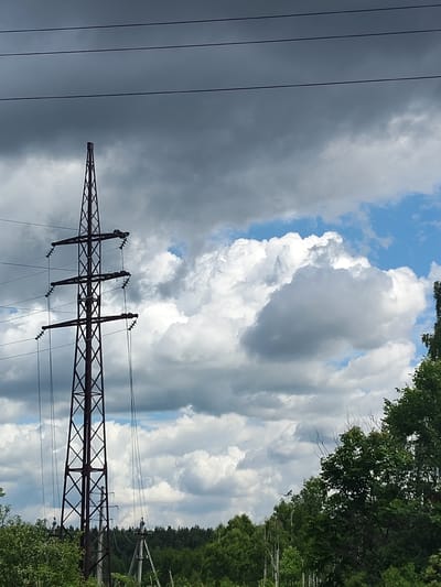 This media file depicts a tall, dark metal power line pylon standing in a rural landscape. The pylon is latticed and supports multiple power lines that stretch across the frame. The sky above is a mix of dramatic, dark grey storm clouds and bright patches of blue sky with fluffy white clouds, suggesting a dynamic weather system. Below the power lines and pylon, a dense line of lush green trees forms the horizon.
To the right of the pylon, the trees are closer to the foreground, their leaves a vibrant green, indicating a warm season, likely summer. The overall setting appears to be a natural, undeveloped area. There is no visible text in the image. The mood conveyed is somewhat dramatic due to the contrasting sky conditions, with the imposing structure of the power line juxtaposed against the natural elements. The time of day is unclear but appears to be daytime. The location context suggests this scene is from Манушкино, Russia.