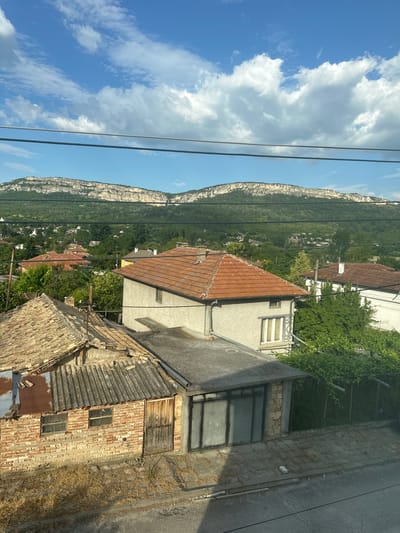 This image captures a scenic view of a village nestled at the base of a dramatic, sheer cliff face. The cliff is a prominent subject, its pale, rocky surface contrasting with the lush green vegetation covering its slopes and the surrounding area. Scattered amongst the greenery are numerous houses with red-tiled roofs, indicative of a European village. In the foreground, closer to the viewer, are several more houses, some appearing older and in a state of disrepair with corrugated roofs and brick walls. Power lines crisscross the upper portion of the image, suggesting the presence of infrastructure. The sky is a vibrant blue, dotted with fluffy white clouds, indicating fair weather. The sunlight suggests it is daytime, likely late morning or early afternoon given the brightness and the shadows. The overall scene evokes a sense of rural tranquility and a close connection to nature. There is no visible text in the image.