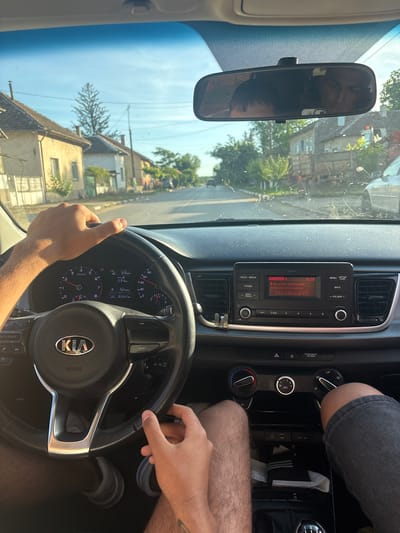 A first-person perspective captures a driver operating a Kia in Hayredin, Bulgaria, as evidenced by the visible houses through the windshield. The driver has both hands on the steering wheel. The dashboard is in clear view, displaying a speedometer reading of 53 km/h and a radio screen showing "BT Audio" with "No artist information" and "No title information." The rearview mirror reflects the driver's face and the street scene behind the vehicle. The image suggests the car is currently in motion.