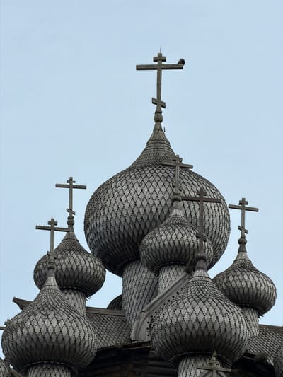This media file captures a close-up, upward-facing view of the ornate wooden domes and crosses of a church, likely the Church of the Transfiguration on Kizhi Island in Russia, given the historical context of Nevskii Parkleskhoz. The intricate construction of the domes is highlighted, with thousands of small wooden shingles creating a scale-like texture. Multiple crosses, also made of wood and featuring traditional Orthodox design elements, are visible atop the domes. One of the crosses has a small bird perched on it.

The sky is overcast and a pale, uniform blue, suggesting either early morning, late afternoon, or a generally cloudy day. There are no people visible in the frame. The focus is entirely on the architectural details of the church structure. The weathered appearance of the wood indicates age and exposure to the elements. The perspective emphasizes the grandeur and complexity of the religious architecture, creating a sense of awe. The image is shot in portrait orientation. There is no visible text within the frame.