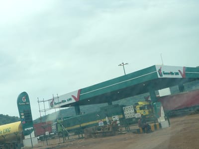 This media file captures a scene at a gas station, likely a liquefied natural gas (LNG) station, in Koton Karifi, Nigeria. The prominent structure is a green and white gas station canopy with the "Greenville LNG" logo visible multiple times. Several large trucks are present, including a yellow tanker truck and a large red trailer truck. One truck is partially covered with a green tarp.
In the foreground, a tall, green vertical sign displays "Greenville" and what appears to be pricing information for different fuel types, with "LNG" also indicated. Two individuals are seated on a low concrete barrier near the sign, one leaning against the other. The ground is dirt and gravel, suggesting a rural or less developed setting.
In the background, lush green hills or mountains rise. The sky is overcast and pale, indicating a cloudy or possibly hazy day. The lighting is diffused, suggesting it might be morning or late afternoon, or simply a heavily overcast day. There's scaffolding erected near the canopy, possibly indicating ongoing construction or maintenance. Visible text includes "Greenville LNG," "LNG," "Entry," and a warning sign that reads "A DANGER NO SMOKING NO OPEN FLAMES NO LIVES." The overall impression is a functional, working gas station in a natural, slightly rugged environment.