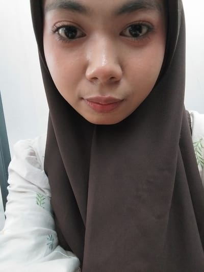 A close-up selfie of a young woman wearing a dark brown hijab and a white shirt with green floral embroidery. She has dark eyes and fair skin. The background is blurred and appears to be indoors. There is no visible indication of the location in Tibojong, Indonesia.