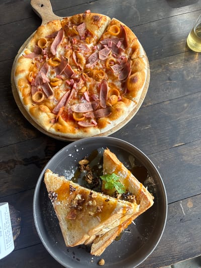 An overhead view captures a dining arrangement on a dark wooden table in Tamiajeng, Indonesia. In the upper half of the frame, a round pizza rests on a light wooden pizza peel. The pizza features a golden-brown crust, melted cheese, a red sauce base, and is topped with numerous irregular strips of light pink processed meat and several ring-shaped processed meat pieces. One slice of the pizza is slightly separated from the main pie. Directly below the pizza, a dark gray ceramic plate holds two triangular pieces of toasted bread or French toast. These are generously drizzled with a translucent golden-brown syrup, topped with a scattering of granola or nut mix, and garnished with a small sprig of fresh mint leaves. Powdered sugar is lightly dusted around the dish on the plate. To the upper right, a clear glass containing a pale yellow liquid is partially visible. In the lower left quadrant, a crumpled white paper receipt, partially legible with text including "Dine In", lies on the wooden table surface. The table itself is composed of dark, horizontal wooden planks.