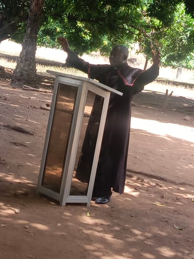 The image shows an older Black man in a dark robe with red trim standing behind a tall, narrow, translucent lectern with a metal frame outdoors in Ninzo South, Nigeria. He has his arms raised. The ground is dirt, and there is a tree with dense green foliage overhead. In the background, there are other trees and possibly a building.