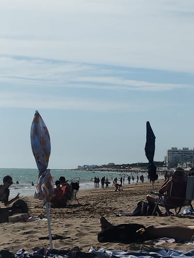 This image depicts a crowded beach scene in Rota, Spain. In the foreground, a person is lying on a towel with their legs extended, partially covered by a dark fabric. Further back, a beach chair is occupied by a person facing away from the camera, with a bag and a towel on the ground nearby. Two closed beach umbrellas are visible, one a pale blue with a patterned design and the other a dark blue. In the mid-ground, numerous people are scattered across the sandy beach and in the shallow water. Some individuals are wading, while others are standing or sitting. In the distance, a line of buildings, likely hotels or apartments, is visible along the coastline, characteristic of the urban development in Rota. The sea is a vibrant turquoise, and the sky is a pale blue with scattered white clouds. The overall impression is a lively summer day at a popular beach resort.