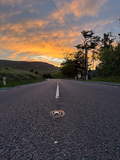 An eye-level shot captures a rural road at sunset in Sconser, United Kingdom, devoid of human presence. The asphalt road, textured with dark gravel, recedes into the distance with a continuous white center line. A metallic road reflector (cat's eye) is embedded in the foreground, slightly right of the center line. White lines delineate the road edges. To the left, a grassy embankment slopes upward towards a range of dark, undulating hills silhouetted against a vibrant orange and yellow sky, indicative of sunset. A white and red striped marker post stands near the road's edge on the left. The sky exhibits dramatic warm hues transitioning to a lighter blue-grey overhead, with scattered, illuminated clouds. On the right, dark green trees and dense foliage border the road, some silhouetted against the sky. A utility pole with an overhead wire is visible on the right side, extending upwards towards the sky. Further down the road on the right, partially obscured by trees and shadows, are several road signs and the silhouette of a building with a visible chimney.