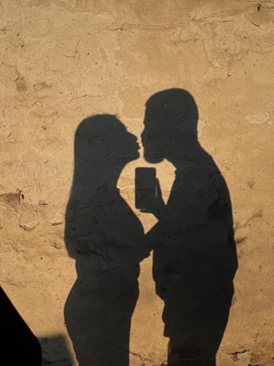 The image shows the shadows of two people, a man and a woman, embracing against a textured, light-colored wall. The couple appears to be taking a selfie, as a shadow of a phone is visible between them. The woman's shadow is on the left, with long hair, and the man's shadow is on the right. The wall exhibits cracks and imperfections, suggesting it is an outdoor surface, possibly in a rustic or historic urban setting. The lighting indicates it is likely late afternoon or early morning, casting long shadows. The location is specified as Ilavche, Ukraine.