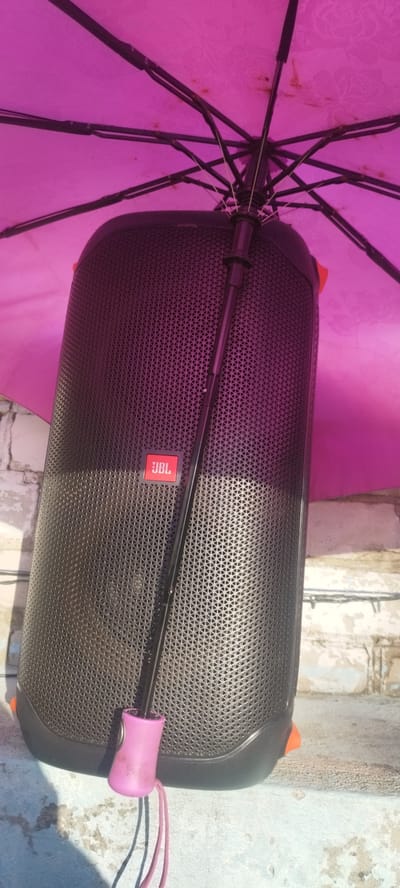 A large, black JBL speaker is positioned upright, partially obscured by a vibrant pink umbrella. The umbrella's metallic ribs and fabric are visible, casting a colorful hue onto the speaker. A black umbrella pole extends down, resting against the speaker's surface, and terminates in a pink, plastic handle with a cord. The speaker itself has a textured, perforated grill and features the distinctive red and white JBL logo. It appears to be outdoors, with a brick wall and a concrete surface visible in the background. Sunlight casts shadows on the concrete, suggesting it is daytime. The setting, combined with the speaker and umbrella, implies a casual outdoor gathering or a setup for portable sound. The overall scene is somewhat rustic, with the brickwork and concrete suggesting an unpolished, possibly rural or urban fringe environment. There is no discernible activity or event taking place; the focus is on the static arrangement of the objects. The image is likely taken in Belarus, based on the provided location context.
